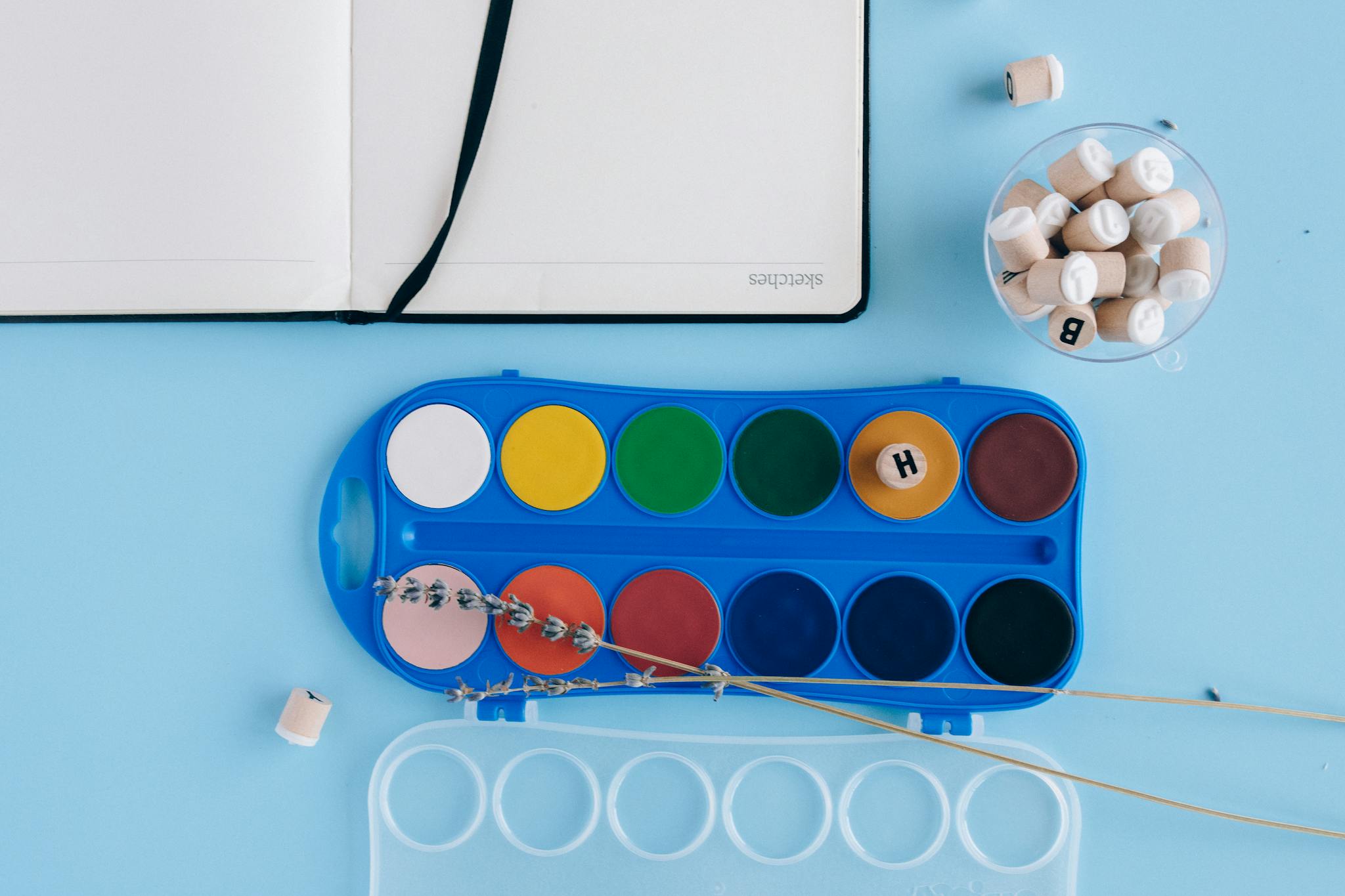 Flat lay of watercolor paints, notebook, and lavender on a blue background.
