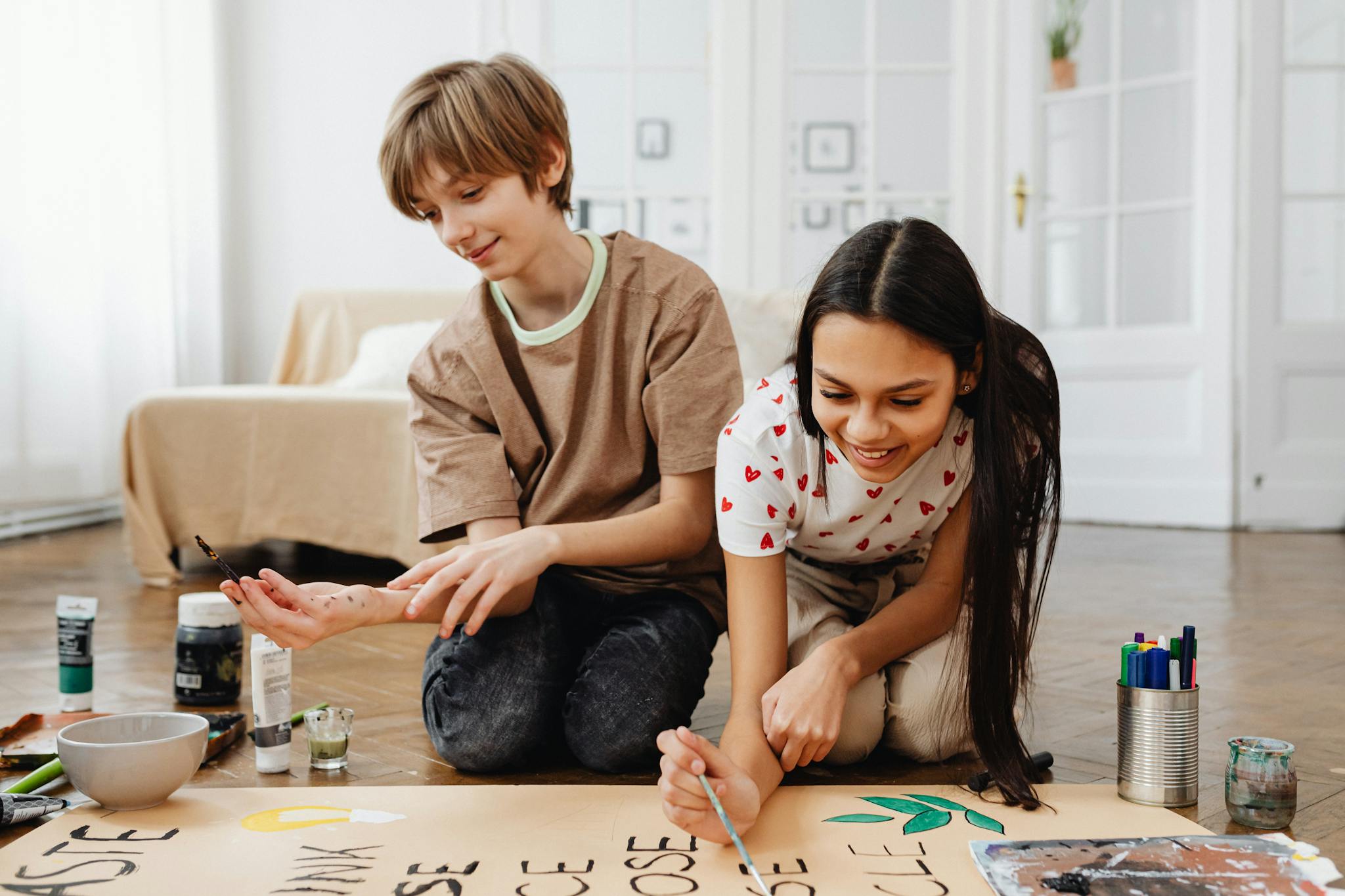 Two teenagers joyfully paint a collaborative school project indoors, fostering creativity and teamwork.
